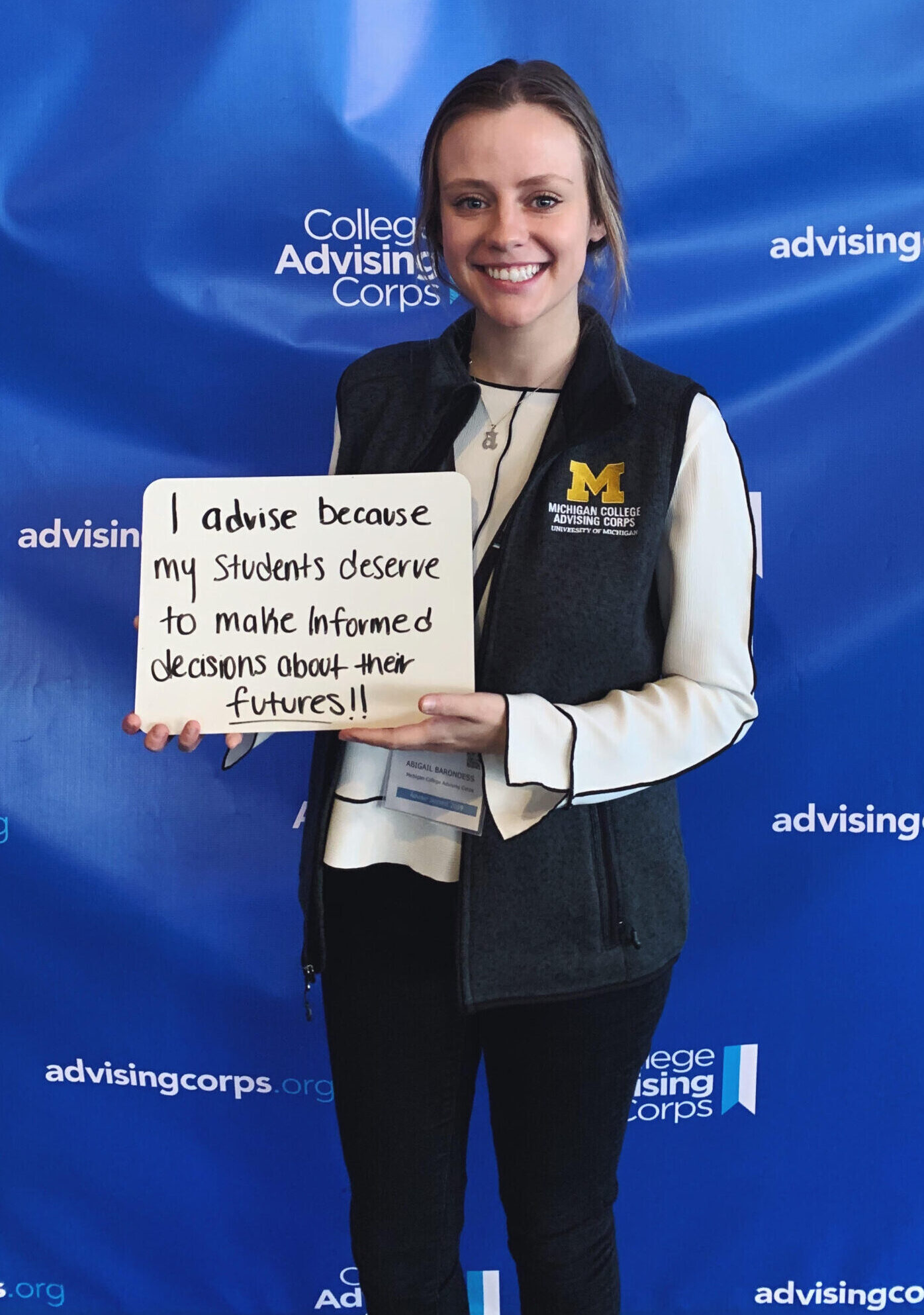 Abigail poses at an event with a handwritten sign that says "I advise because my students deserve to make informed decisions about their futures!"